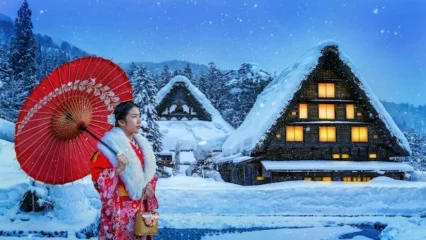 Woman in traditional Japanese winter kimono with red umbrella at snow-covered Shirakawa-go village, wearing furisode with white fur collar against traditional gassho-zukuri farmhouses
