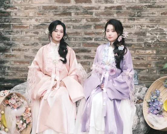 Two women dressed in traditional Korean hanbok sitting side by side against a weathered brick wall — one wearing a soft pink jeogori with sheer overlay and white chima, the other in a lavender purple jeogori with light blue embroidery and white chima, both styled with floral hair accessories, surrounded by woven baskets filled with pink and purple flowers
