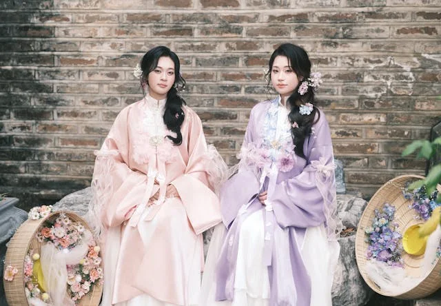 Two women dressed in traditional Korean hanbok sitting side by side against a weathered brick wall — one wearing a soft pink jeogori with sheer overlay and white chima, the other in a lavender purple jeogori with light blue embroidery and white chima, both styled with floral hair accessories, surrounded by woven baskets filled with pink and purple flowers