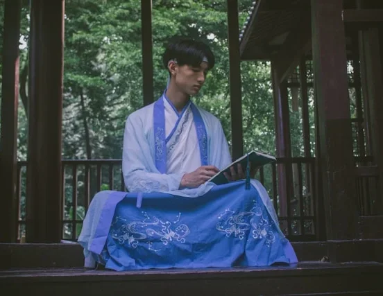 A man wearing korean traditional clothing for men in white and blue hanbok, sitting outdoors in a wooden pavilion surrounded by greenery