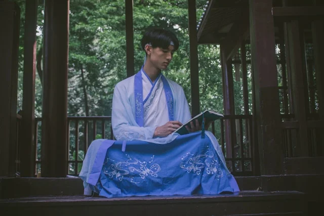 A man wearing korean traditional clothing for men in white and blue hanbok, sitting outdoors in a wooden pavilion surrounded by greenery