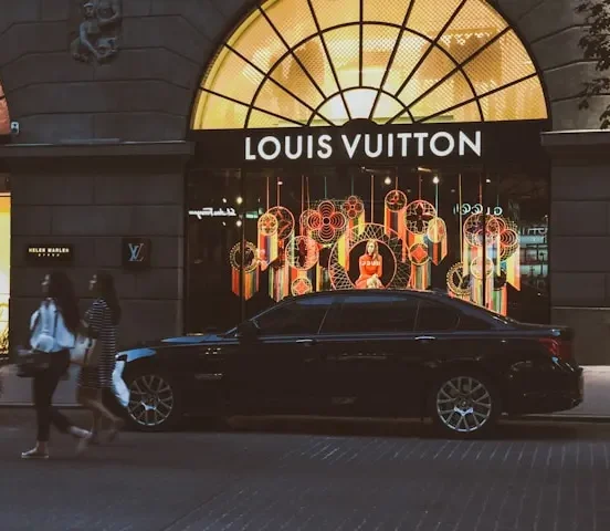 Nighttime street view of a Louis Vuitton storefront with illuminated window displays, a black sedan parked in front, and pedestrians walking past the entrance.