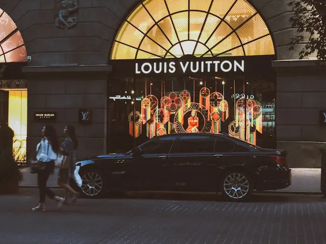 Nighttime street view of a Louis Vuitton storefront with illuminated window displays, a black sedan parked in front, and pedestrians walking past the entrance.