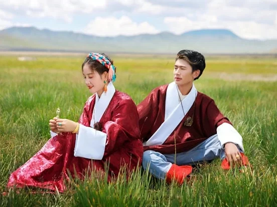 A young woman and man sitting back-to-back in a green meadow wearing matching deep red traditional robes with white inner collars and light blue trousers, with mountains visible in the background