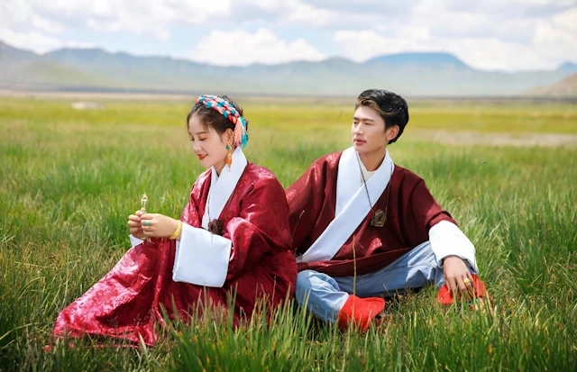 A young woman and man sitting back-to-back in a green meadow wearing matching deep red traditional robes with white inner collars and light blue trousers, with mountains visible in the background