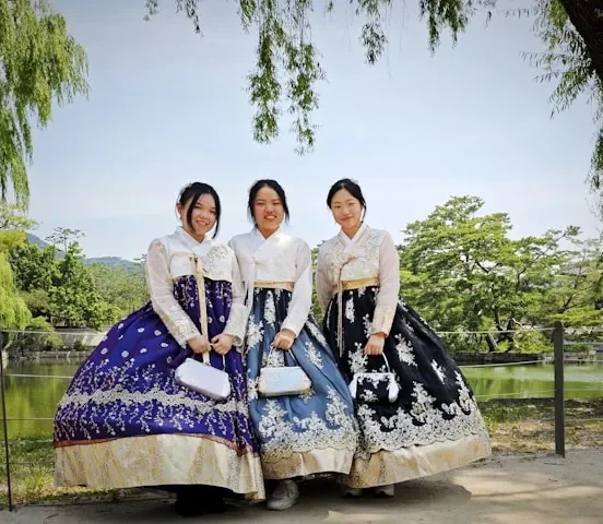 Three young women wearing traditional Korean hanbok — white jeogori jackets paired with richly embroidered chima skirts in purple, light blue, and black — posing together outdoors near a pond surrounded by willow trees