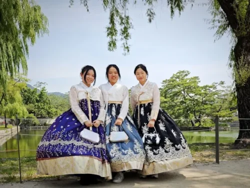 Three young women wearing traditional Korean hanbok — white jeogori jackets paired with richly embroidered chima skirts in purple, light blue, and black — posing together outdoors near a pond surrounded by willow trees
