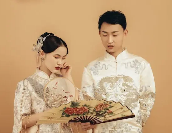 Elegant couple in traditional embroidered attire holding decorative fans during a cultural wedding photoshoot against a beige background.