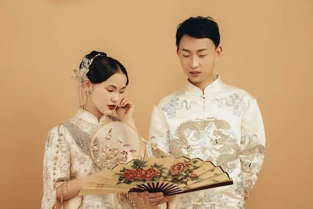 Elegant couple in traditional embroidered attire holding decorative fans during a cultural wedding photoshoot against a beige background.