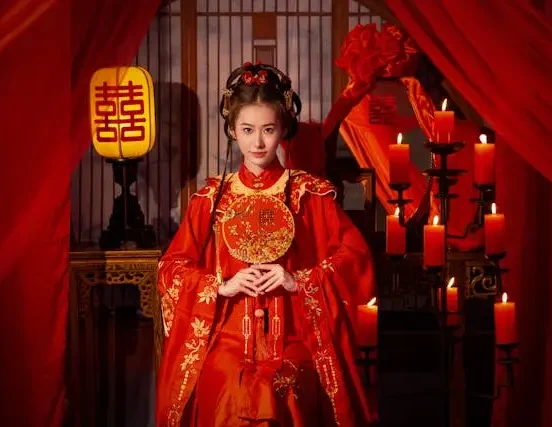 Woman wearing traditional Chinese wedding attire in a red decorated room with candles and cultural decorations