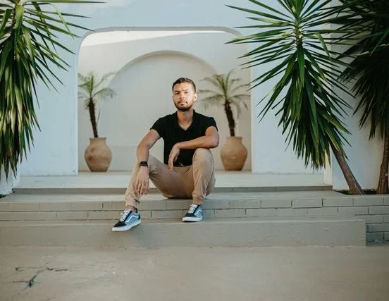 A man in a black V-neck T-shirt and beige chinos sits casually on outdoor concrete steps between tropical plants, wearing black-and-white sneakers in a modern minimalist setting with white archway architecture in the background.
