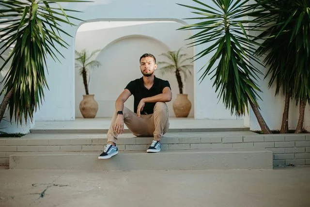A man in a black V-neck T-shirt and beige chinos sits casually on outdoor concrete steps between tropical plants, wearing black-and-white sneakers in a modern minimalist setting with white archway architecture in the background.