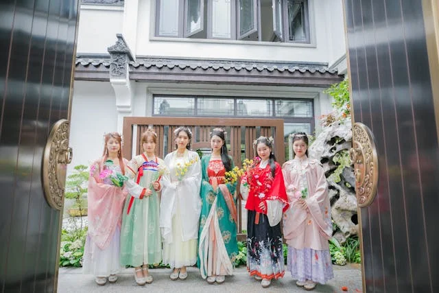 Group of women wearing traditional Chinese Hanfu dresses standing at the entrance of a traditional-style building with floral decorations.