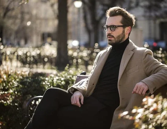 Stylish man wearing a beige overcoat and black turtleneck while sitting on a park bench in an urban setting during daylight.