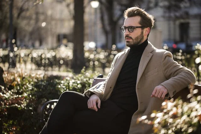 Stylish man wearing a beige overcoat and black turtleneck while sitting on a park bench in an urban setting during daylight.