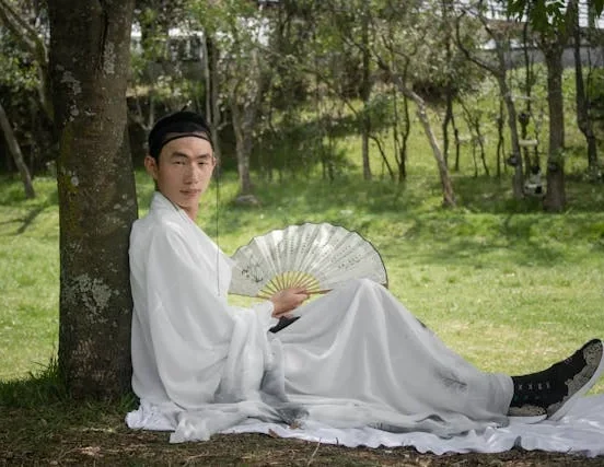 A man wearing flowing white traditional East Asian robes sits beneath a tree in a grassy park, holding an open folding fan while relaxing in the shade.