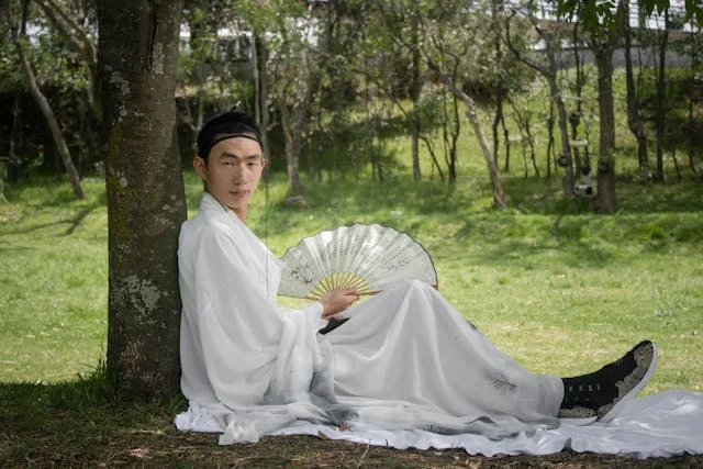 A man wearing flowing white traditional East Asian robes sits beneath a tree in a grassy park, holding an open folding fan while relaxing in the shade.