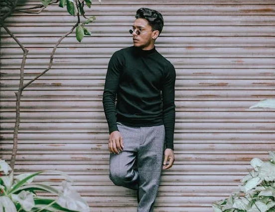 Man wearing a black turtleneck sweater and gray trousers posing casually in front of a metal shutter with plants surrounding the scene.