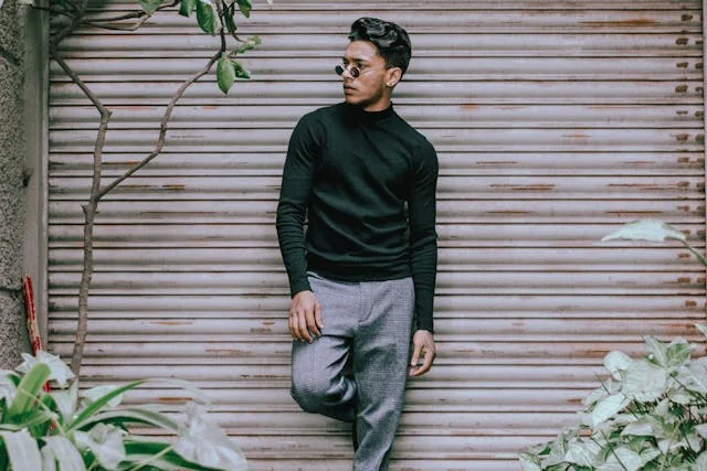 Man wearing a black turtleneck sweater and gray trousers posing casually in front of a metal shutter with plants surrounding the scene.