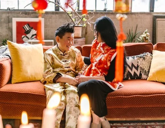 Two people wearing traditional festive clothing sit together on a couch surrounded by candles and red hanging decorations during a holiday celebration.