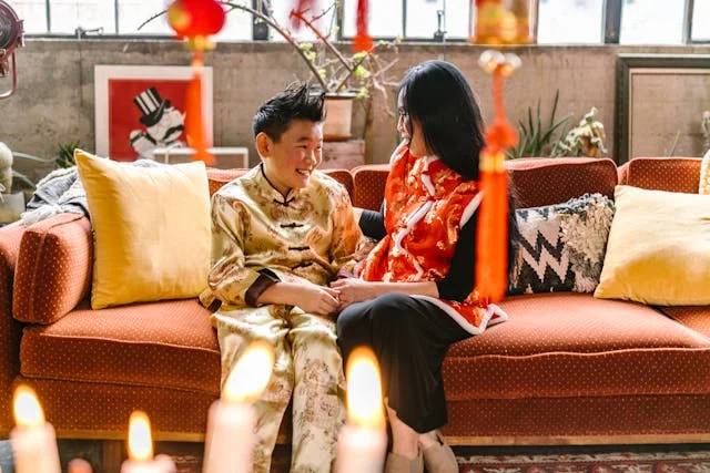 Two people wearing traditional festive clothing sit together on a couch surrounded by candles and red hanging decorations during a holiday celebration.