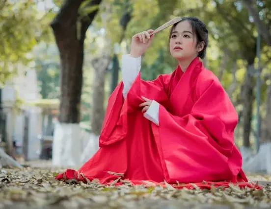 Young woman wearing traditional Korean clothing for women — a flowing red chima skirt and white jeogori jacket — sitting outdoors among autumn leaves holding a folding fan