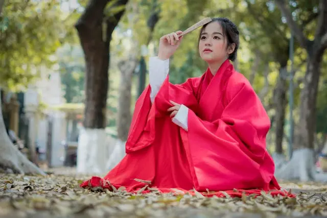 Young woman wearing traditional Korean clothing for women — a flowing red chima skirt and white jeogori jacket — sitting outdoors among autumn leaves holding a folding fan